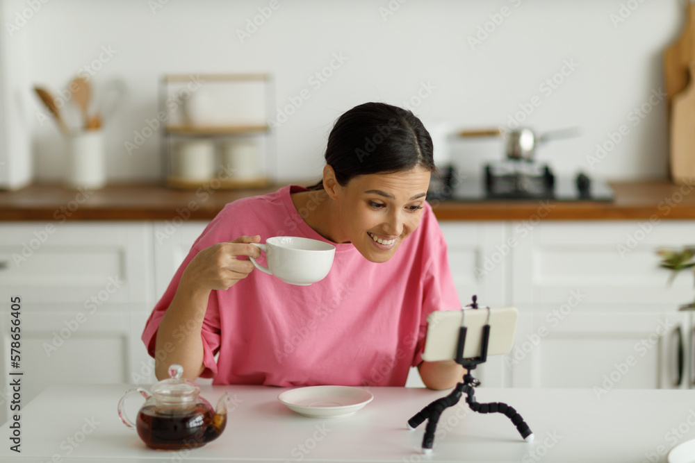 Attractive smiling young brunette woman in pink oversize T-shirt is drinking tea in kitchen. Happy pretty girl with snow-white smile greets friend, communicating by video call online on mobile phone. Attractive smiling young brunette woman in pink oversize T-shirt is drinking tea in kitchen. Happy pretty girl with snow-white smile greets friend, communicating by video call online on mobile phone.