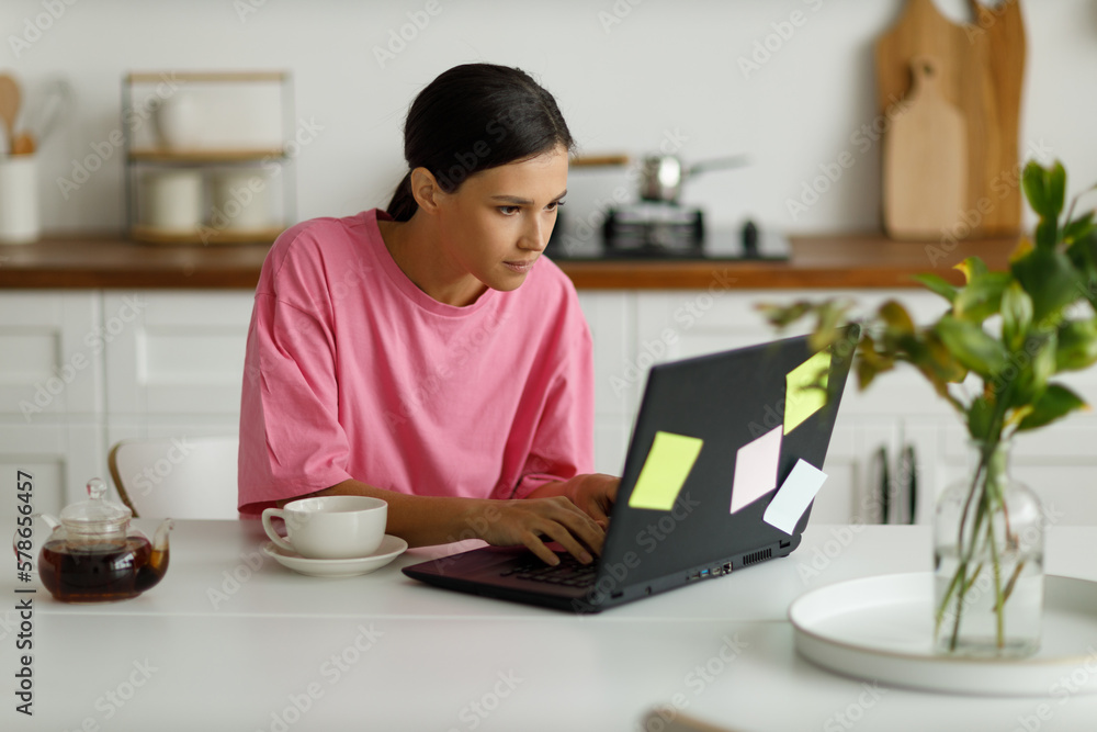 Happy girl with laptop checks her mail, looks through messages or funny videos. Video communication online. Beautiful young woman works remotely from home, sitting at the dining table in the kitchen. Happy girl with laptop checks her mail, looks through messages or funny videos. Video communication online. Beautiful young woman works remotely from home, sitting at the dining table in the kitchen.