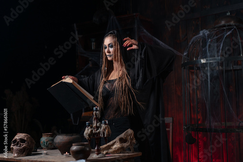 Halloween concept. Witch dressed black hood with dreadlocks standing dark dungeon room use magic book for conjuring magic spell