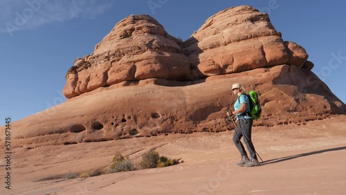 Hiker mature woman with backpack and trekking poles hiking at flat and smooth rock monolith of orange sandstone. Walking background rock formations in Arches national park Utah. Slow motion side view photo