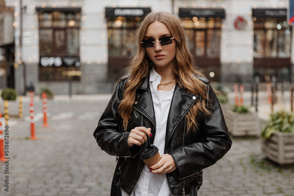 Fashionable  blonde woman model with  black leather jacket, style sunglasses  and coffee walking the city street