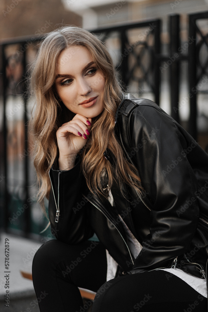 Close up portrait of a beautiful smiling young blonde woman in a black leather jacket on the  a city street background