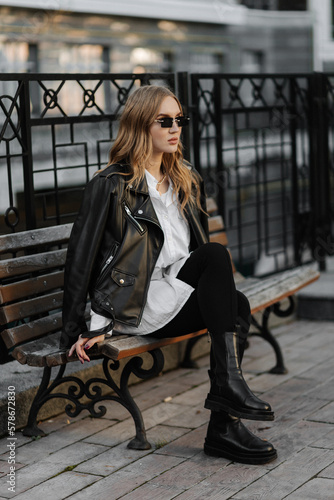 Fashionable  blonde woman model with  black leather jacket and style sunglasses sitting on a bench at the city 