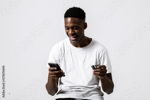 Happy delighted african american guy student received first sallary holding credit card and cellphone in hands payment process making purchase online over grey background in studio isolated. photo
