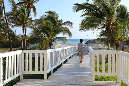 Picturesque view to wooden path to tropical beach with palm trees. Lonely woman in dress walking towards ocean coast, tourist resort on island