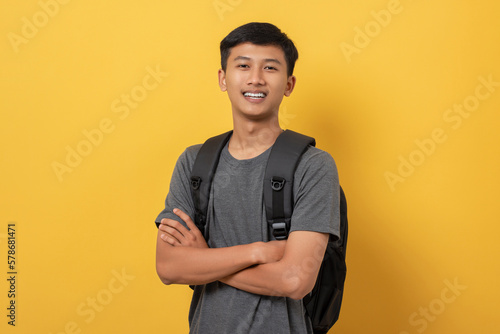 Portrait of smiling confident young college student with books and backpack isolated on yellow background