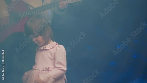 Little girl on stage in pink vintage dress, she dances, her father plays acoustic guitar. Color music is shining and smoke is billowing. Father Day. Performance on stage. Hobbies and creativity