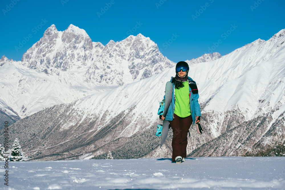 Snowboarder female walking with snowboard in beautiful mountain peaks covered with snow on background