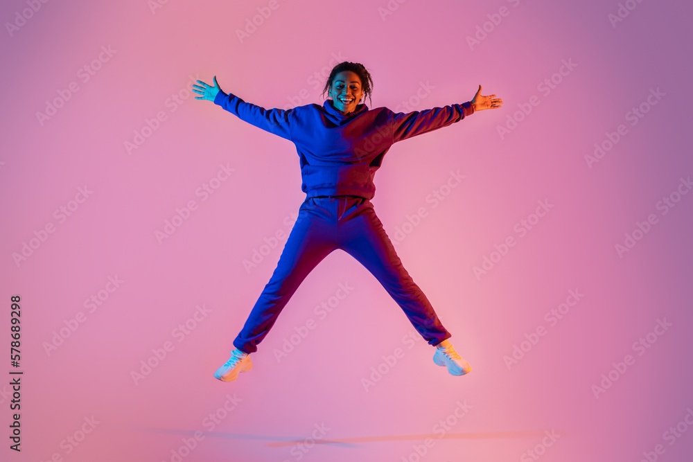 Full body length of sporty african american young woman jumping up in star shape, having fun over pink neon background