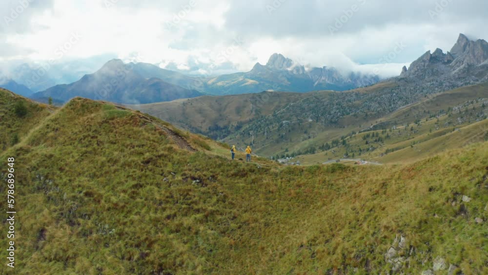 Tourists stand on hill top exploring high rocky cliffs around Pass Giau. Hikers examine mountain nature standing on pathway facing valley under cloudy sky Tourists stand on hill top exploring high rocky cliffs around Pass Giau. Hikers examine mountain nature standing on pathway facing valley under cloudy sky