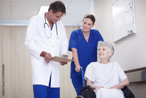 You can rest easy. A female nurse pushing a senior patient in a wheelchair down the corridor while talking with her doctor.