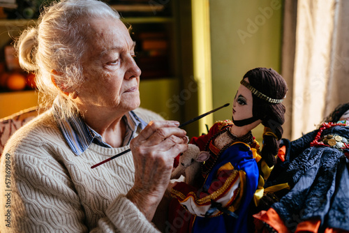 Old Caucasian beautiful grey haired artist woman sewing doll at home. Maker exclusive colorful handmade needlework.