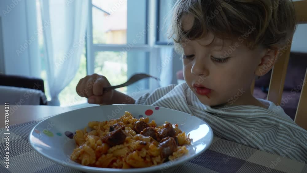Child eating pasta food for lunch at home. Authentic lifestyle scene of kid eats noodles by himself with fork Child eating pasta food for lunch at home. Authentic lifestyle scene of kid eats noodles by himself with fork