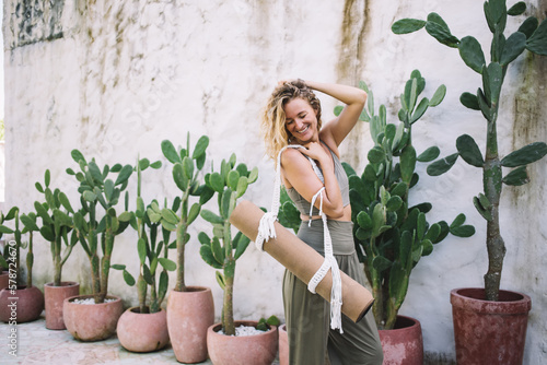 Smiling woman with yoga mat standing near green plants