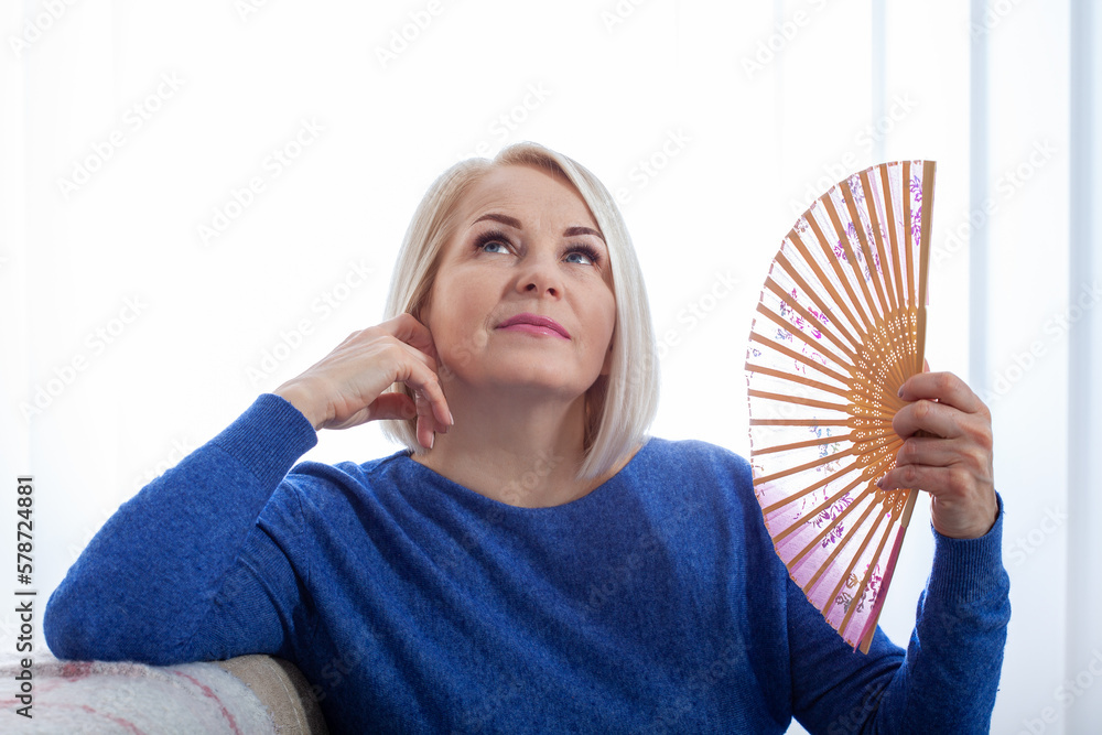 Mature woman experiencing hot flush from menopause. A woman in her fifties sitinds fanning herself with a handheld fan, her face flush with the heat of menopause. - obrazy, fototapety, plakaty 