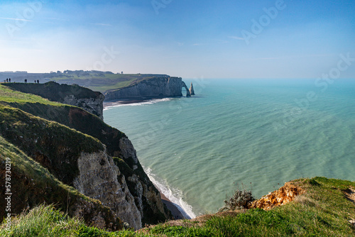 Etretat Cliffs, A cliff with a large arch in the ocean, A group of people on a cliff overlooking a body of water, A cliff with a body of water and a rock formation, A blue sky with clouds and a hill