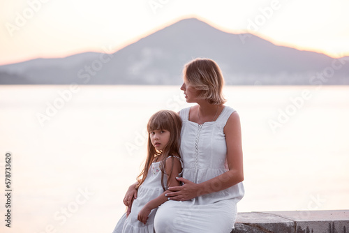 Pregnant Millennial Mother sit with daughter at sunset by sea. Middle aged woman hugs little girl