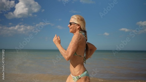 Happy athletic mature senior woman jogging on beach wearing bikini and sunglasses. Adventure is ageless. © Robert Peak