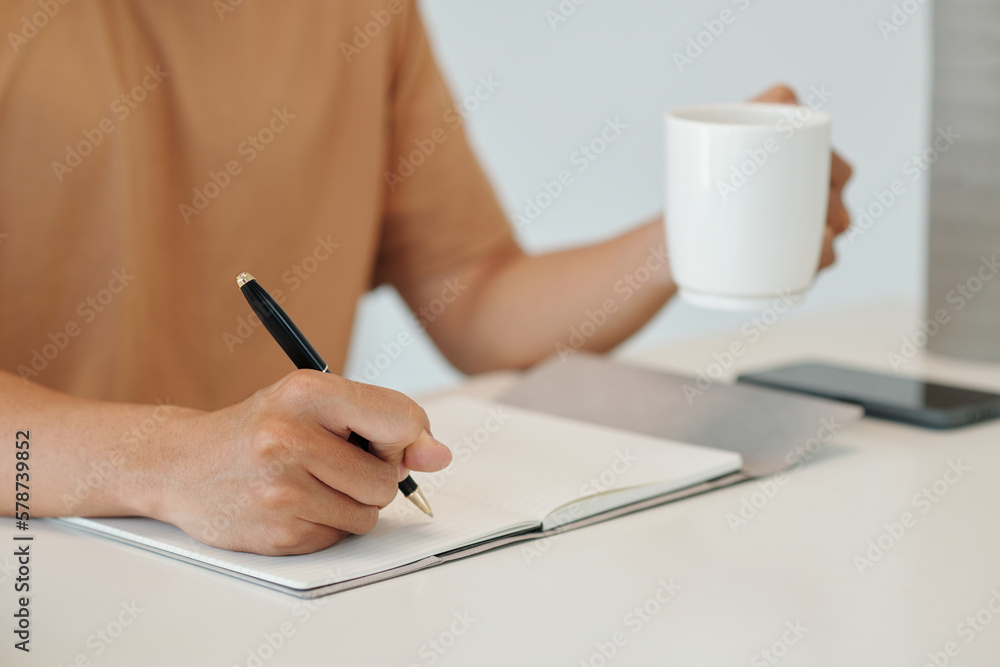 Closeup image of young man drinking cup of morning coffee and writing down plans for the day Closeup image of young man drinking cup of morning coffee and writing down plans for the day