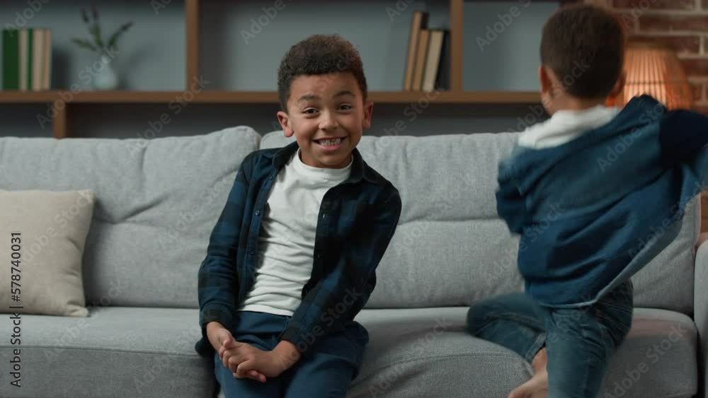 Funny little african american child kid boy son on couch at home smiling sibling brother junior jump on sofa shout. Two ethnic siblings children boys kids together portrait in living room waving hello Funny little african american child kid boy son on couch at home smiling sibling brother junior jump on sofa shout. Two ethnic siblings children boys kids together portrait in living room waving hello