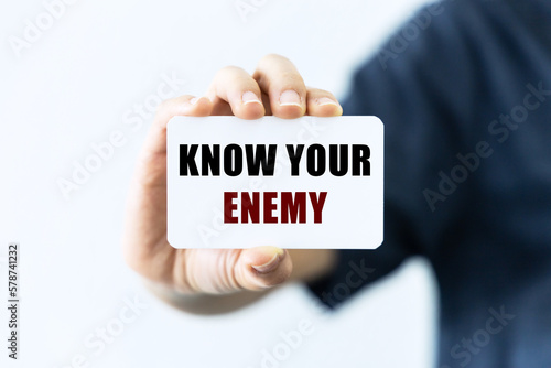 Know your enemy text on blank business card being held by a woman's hand with blurred background. Business concept about knowing opponents.