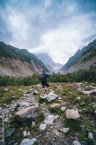 Traveler man with a backpack standing on a stone, On the way, walk the track. A valley among majestic high mountains.. Back view. The Chalaadi Glacier, Georgia, Caucasus. Vertical photo.