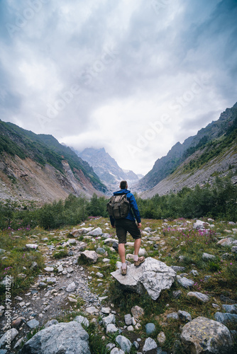 Traveler man with a backpack standing on a stone, On the way, walk the track. A valley among majestic high mountains.. Back view. The Chalaadi Glacier, Georgia, Caucasus. Vertical photo. photo