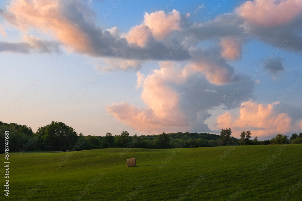 Fototapeta premium Landscape with hay bale at freshly mown vast field at sunset hour. Sunny day, blue sky, soft light. Farming, agriculture, harvest, pasture, livestock concept. 