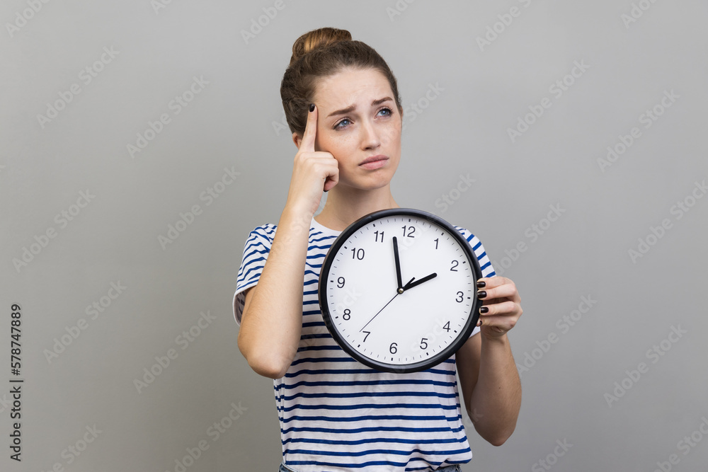 Pensive thoughtful woman wearing striped T-shirt holding wall clock, thinking about deadline. Pensive thoughtful woman wearing striped T-shirt holding wall clock, thinking about deadline.