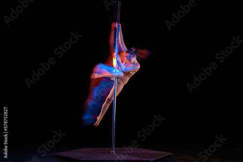 Acrobatic dance. Young girl performing pole dance isolated over black studio background with mixed neon lights. Concept of sport and dance, beauty of movements, action, modern style