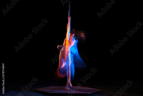 Passionate hobby. Young artistic girl performing pole dance isolated over black studio background with mixed neon lights. Concept of sport and dance, beauty of movements, action, modern style