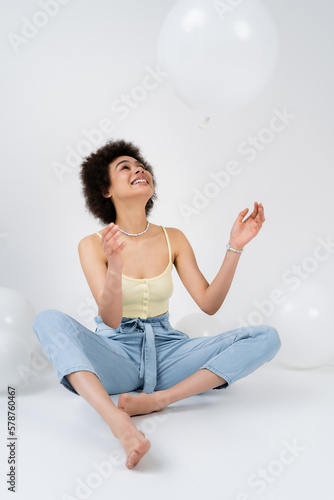 Positive african american woman throwing balloon on grey background.