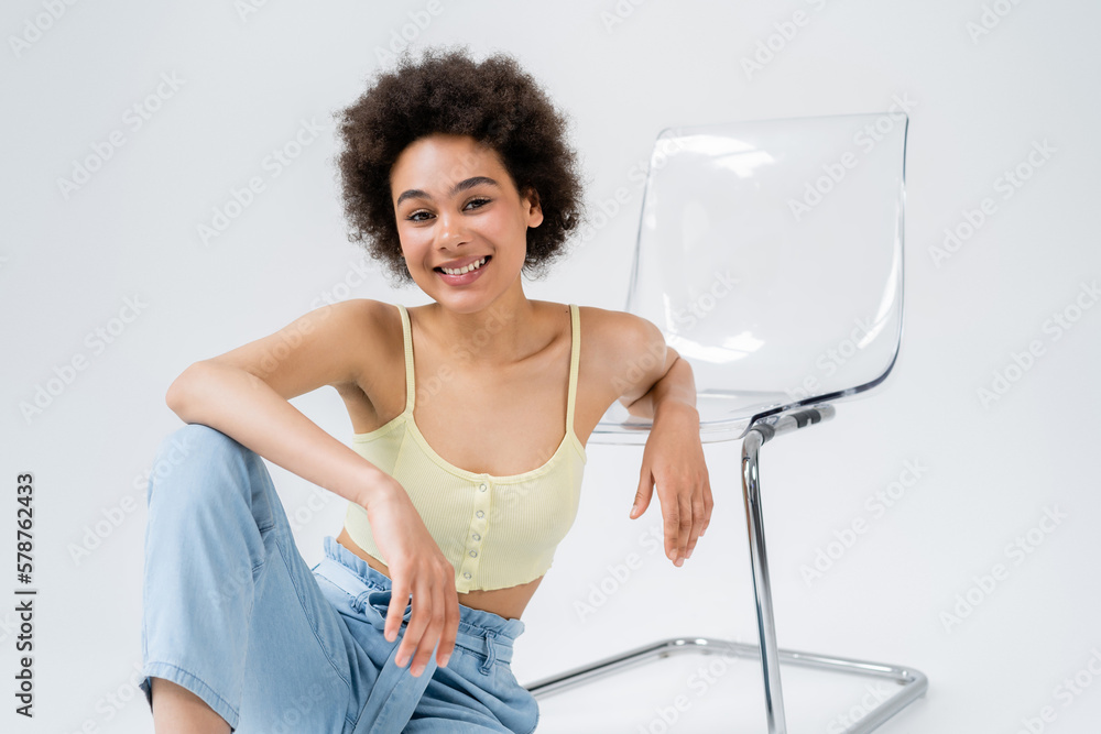 Curly african american woman sitting near modern chair on grey background. Curly african american woman sitting near modern chair on grey background.