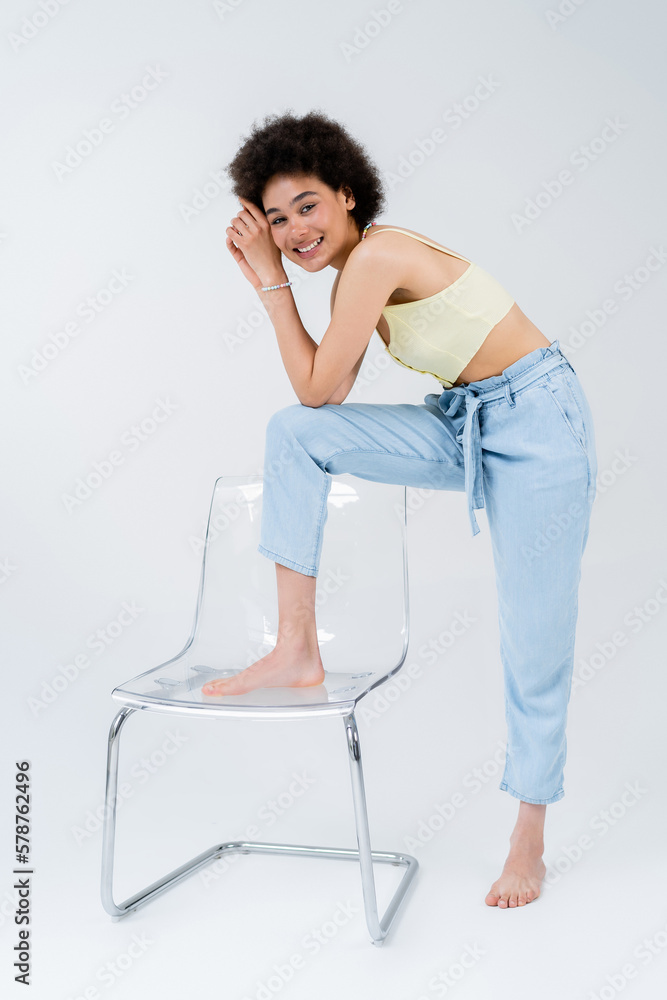 Full length of african american woman posing near modern chair on grey background.