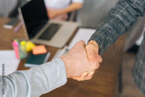 Close-up cropped shot of unrecognizable business partners handshaking after close deal and signed agreement at office over table with laptop and paper contract. Success, dealing, greeting concept.
