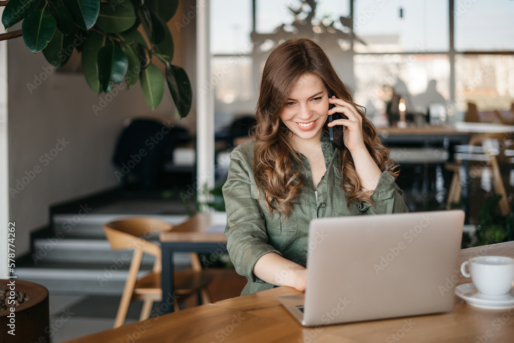Woman books tickets for concert. Portrait of good-looking caucasian girl sitting in cafe, drinking coffee, talking on smartphone, looking at laptop screen with broad smile, checking profile of friend Woman books tickets for concert. Portrait of good-looking caucasian girl sitting in cafe, drinking coffee, talking on smartphone, looking at laptop screen with broad smile, checking profile of friend