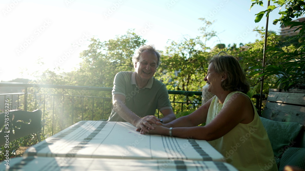 A senior couple authentic real life laugh and smile together. Happy candid married mature husband and wife holding hands sitting outdoors in home patio backyard