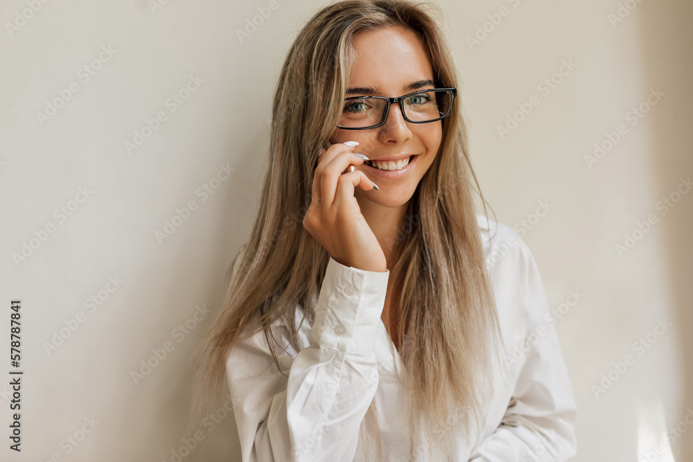 Spectacular charming woman with loose hair and happy smile wearing glasses and white shirt is posing to camera over isolated background. Indoor photo of glamorous happy girl Spectacular charming woman with loose hair and happy smile wearing glasses and white shirt is posing to camera over isolated background. Indoor photo of glamorous happy girl
