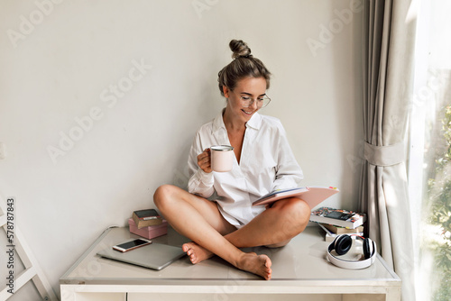 Nice stylish caucasian woman posing looking at camera sitting on the table with legs crossed on the table in white room. Blond wears white shirt with coffee and notebook. Beauty and youth concept