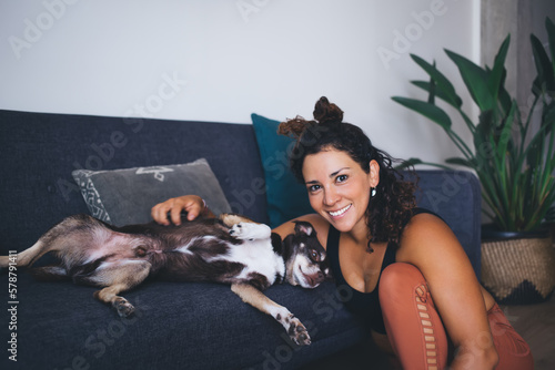 Funny Caucasian female cynologist resting in home living room and smiling at camera during leisure with mongrel dog from shelter, portrait of joyful woman with pet best friend enjoying weekend photo