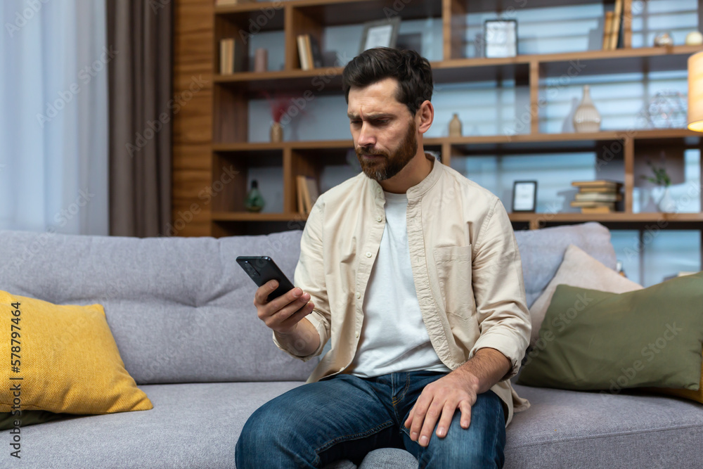 Communication problems. A serious young man in a beige shirt sits on the sofa at home, holds the phone, looks worriedly at the screen, types a message.