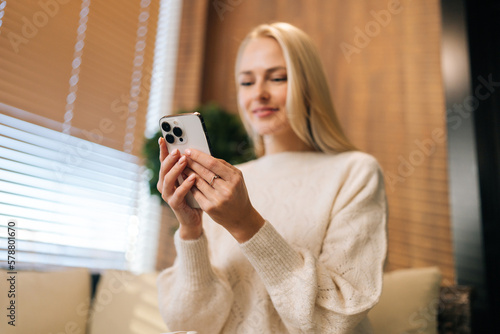 Low-angle view of pretty millennial blonde woman looking at mobile phone screen watching funny photo or video, enjoying pleasant distant communication in social network, sitting on sofa by window.