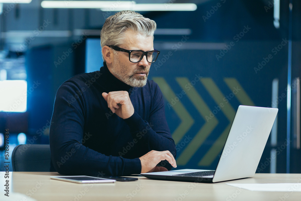 Thinking businessman inside the office with a laptop, mature adult boss thinking about the decision for working sitting with a computer. Thinking businessman inside the office with a laptop, mature adult boss thinking about the decision for working sitting with a computer.