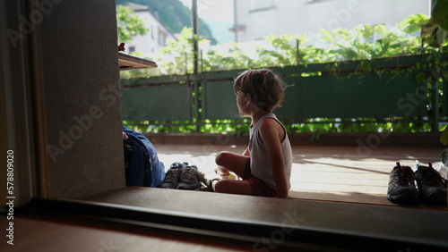 Child putting shoe by doorstep home entrance. Kid sitting on floor puts tennis shoes