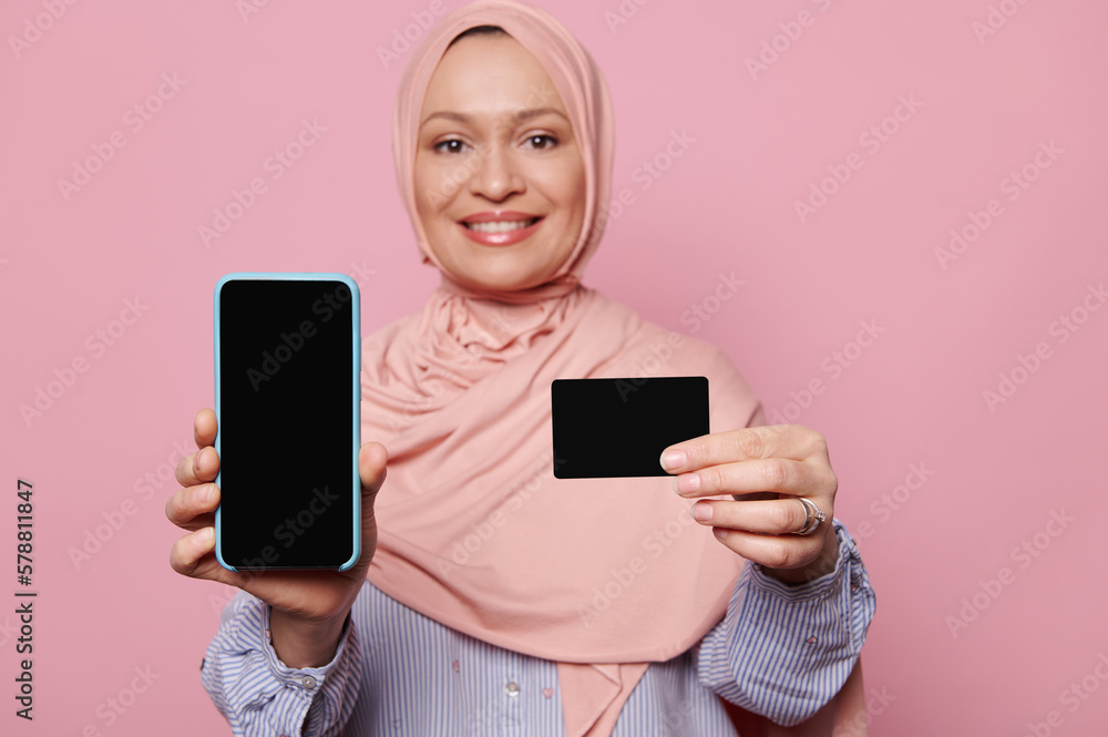 Selective focus on a smartphone with black blank screen and a plastic credit card, in the hands of a blurred smiling Arab Muslim pretty woman, showing a free space for ads, isolated on pink background Selective focus on a smartphone with black blank screen and a plastic credit card, in the hands of a blurred smiling Arab Muslim pretty woman, showing a free space for ads, isolated on pink background