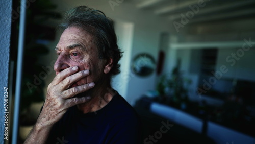 A fearful senior man standing by window with contemplative expression. Anxious expression of a mature older man in 70s at home indoors. apprehensive emotion