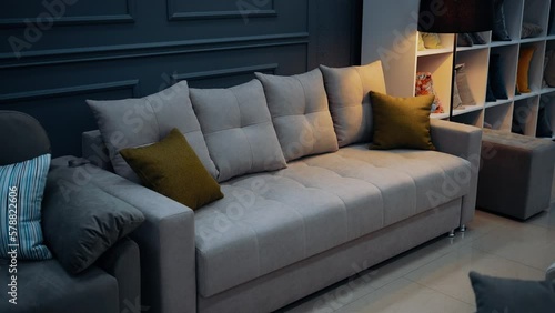 Beige sofa, stylish with four large beige pillows, two small yellow pillows. Decorative lamp next to a large shelf with colorful cushions.
