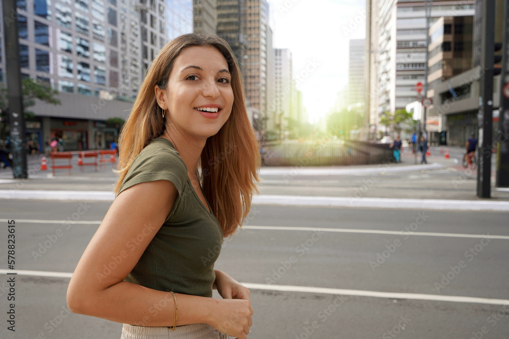 Fototapeta premium Brazilian self-confident young woman smiling at camera with Paulista Avenue on the background in the morning, Sao Paulo, Brazil