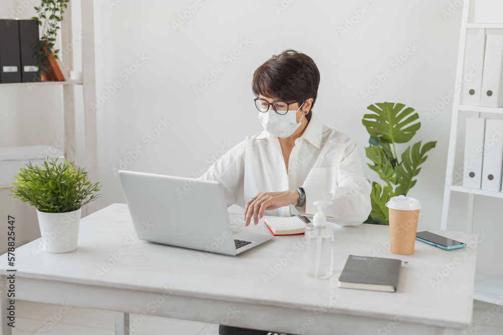 Attractive mature business woman working on laptop in her workstation in medical face mask - manager office and pandemic covid-19 concept