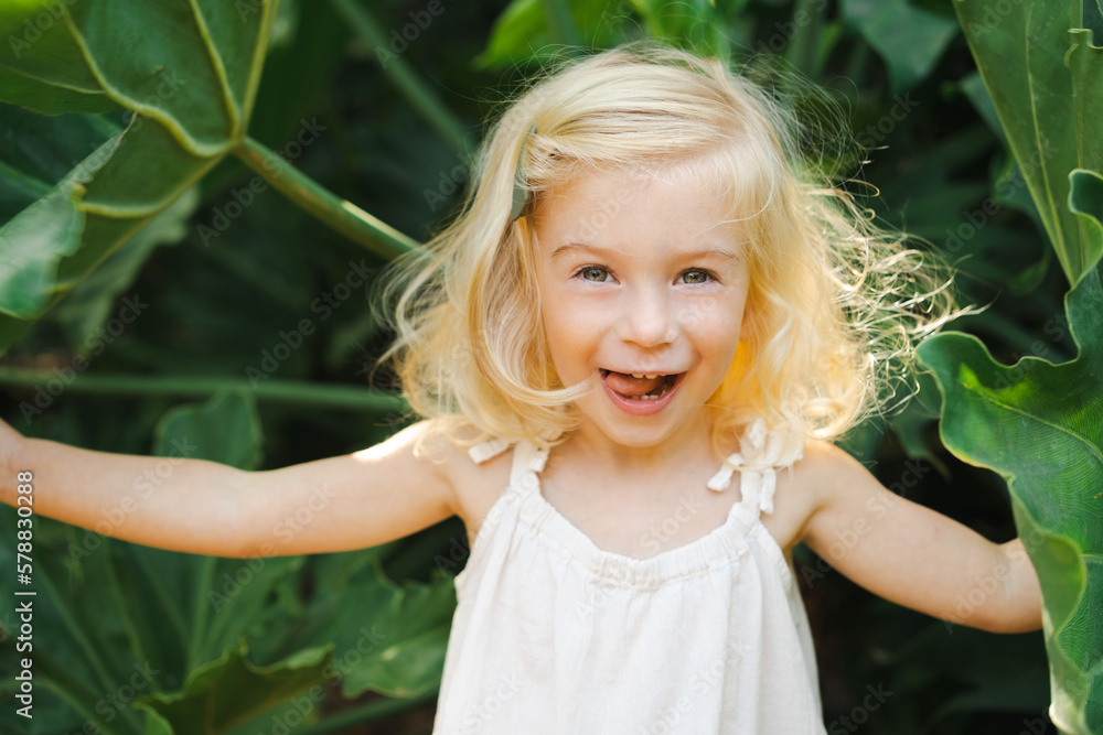 Cute caucasian pretty blonde two year old girl, toddler, kid among big green exotic plants,leaves smiling looking at camera. Natural beauty,perfect skin, health,cosmetics concept.Close up shot. Cute caucasian pretty blonde two year old girl, toddler, kid among big green exotic plants,leaves smiling looking at camera. Natural beauty,perfect skin, health,cosmetics concept.Close up shot.